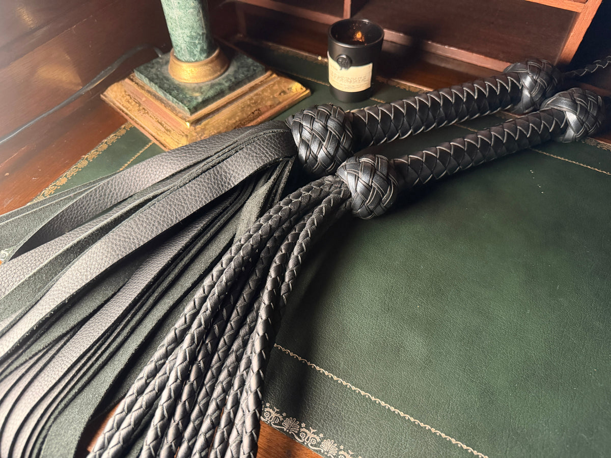 Black leather floggers coiled on a green leather desk, The Garrison by Collared Foxy