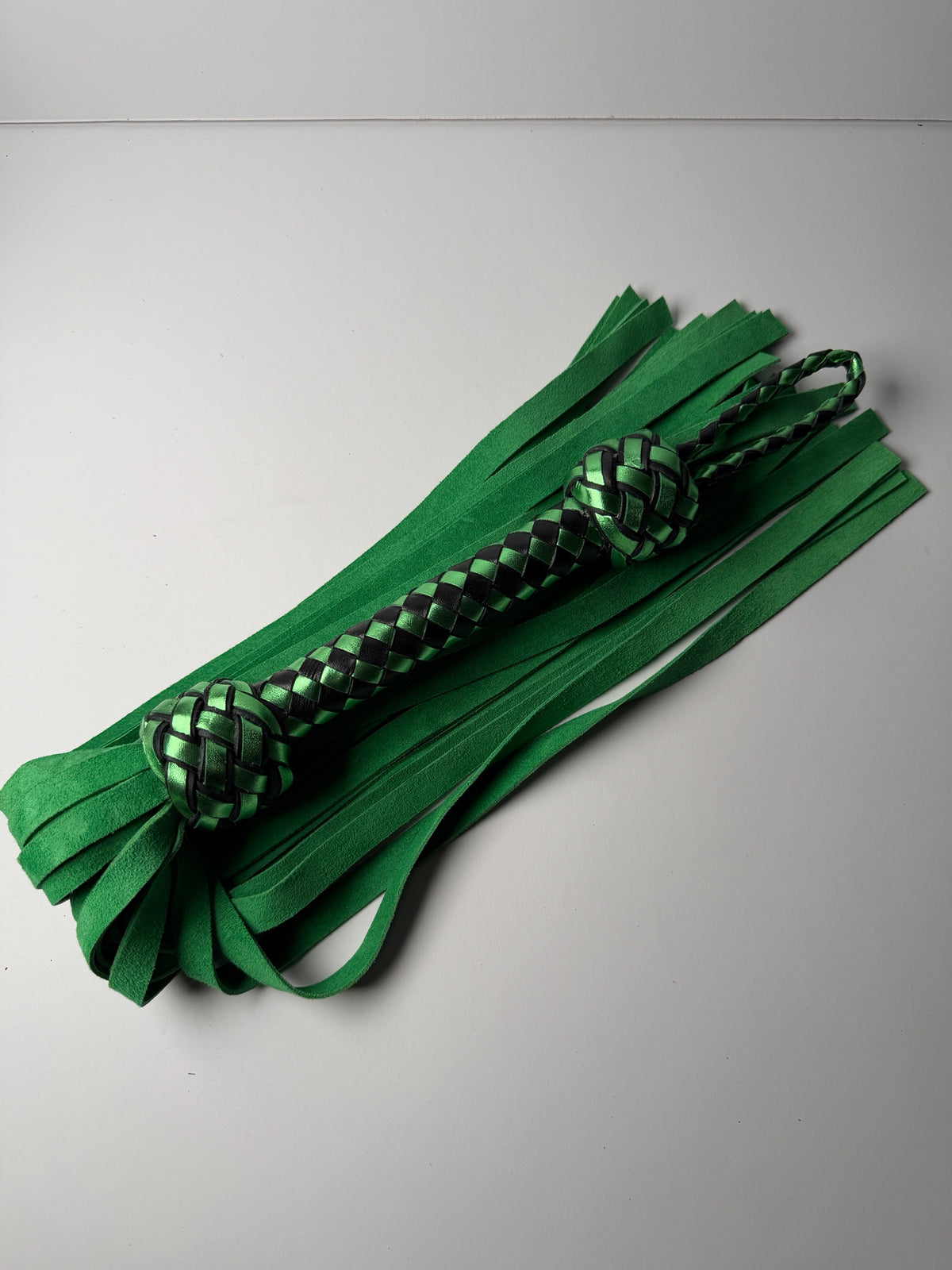 Peridot flogger laid flat, green cowhide suede falls spread beneath the metallic green and black braided handle